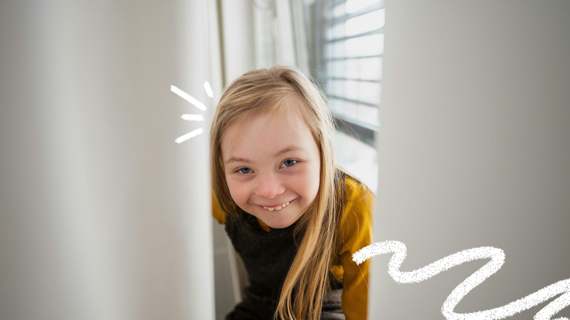 Menina com síndrome de Down sorri enquanto espia por entre uma cortina branca, vestindo blusa amarela e moletom escuro.