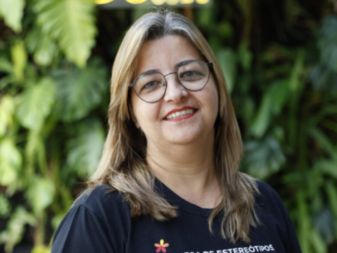 Mulher de óculos e cabelo loiro escuro sorri em frente a plantas, usando camiseta preta com frase sobre inclusão, com letreiro do Mano Down ao fundo.