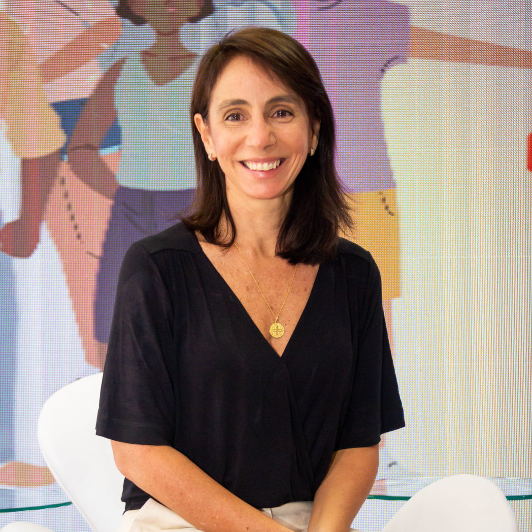 Retrato de Gabriella Bighetti sentada em uma cadeira, sorrindo para a cùmera. Ela veste blusa preta e calça clara, com um colar dourado. Ao fundo, hå um painel com ilustraçÔes de pessoas, sugerindo um ambiente institucional ou de evento ligado a causas sociais.