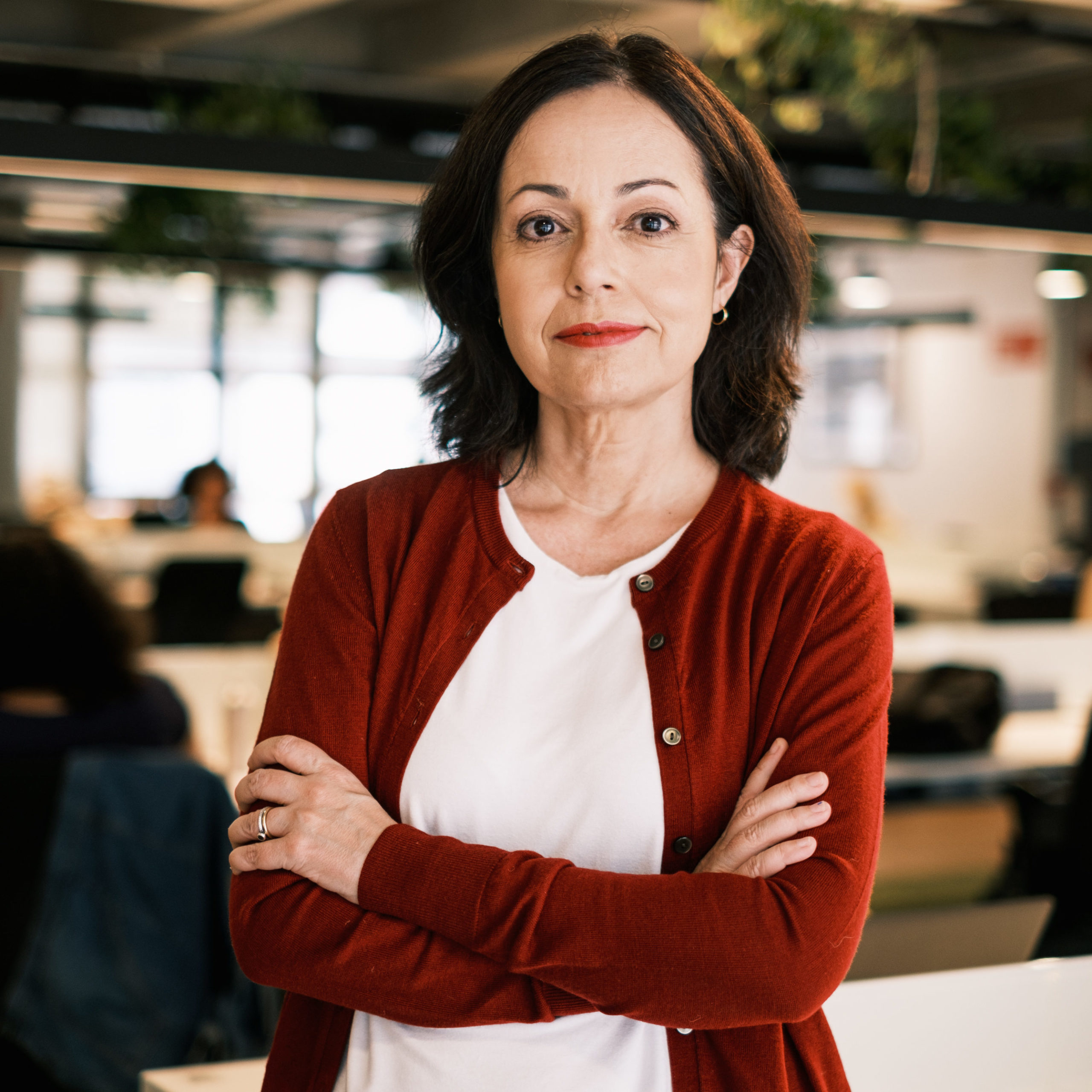 Retrato de Patricia Mota Guedes em um escritório. Ela estå de braços cruzados, vestindo camiseta branca e casaco vermelho, com expressão confiante. Ao fundo, hå um espaço de trabalho com mesas, cadeiras e pessoas desfocadas, sugerindo um ambiente corporativo.