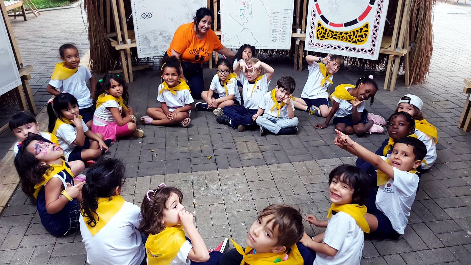Um grupo de crianças com uniforme escolar e bandanas amarelas amarradas no pescoço estão sentadas em roda e posam para a foto junto da professora Edna Monteiro, que usa camiseta laranja com a palavra "criança".