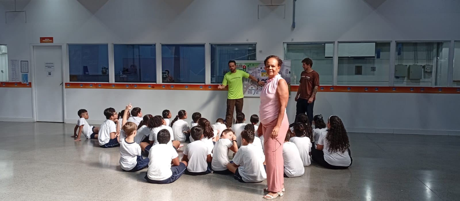 A professora Maria Ester Pereira Moreira de Camillo é uma mulher negra que veste calça e blusa rosa. Ela está de pé segurando um óculos em frente a um grupo de crianças sentadas no chão e de costas para a foto.