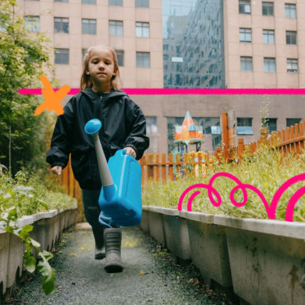 Criança caminha por um canteiro verde em área urbana, segurando um regador azul, entre plantas e cercas de madeira, com prédios ao fundo, em um espaço que mistura natureza e cidade.