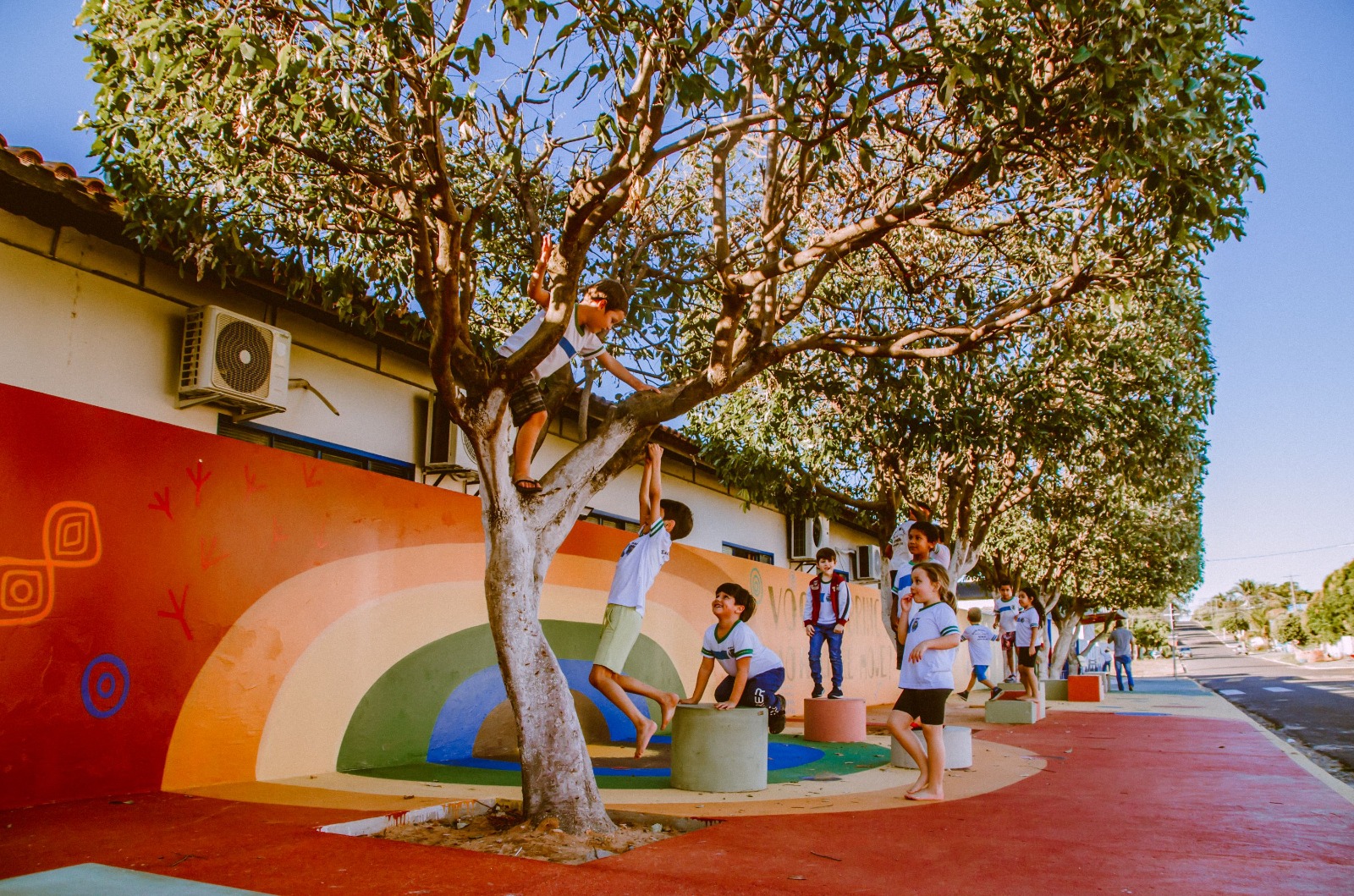 Crianças brincam ao ar livre em um espaço urbano colorido, com árvores alinhadas na calçada; uma delas sobe em um galho enquanto outras observam e interagem, em área com pintura de arco-íris no chão e muro ao fundo.