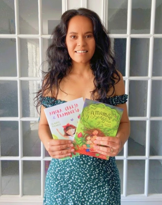 Mulher sorrindo para a câmera, usando vestido estampado e segurando dois livros infantis nas mãos, diante de uma porta de vidro branca.