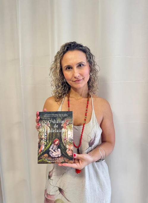 Mulher em pé diante de uma cortina clara, segurando um livro à frente do corpo e olhando para a câmera. Usa vestido claro e colar de contas vermelhas.