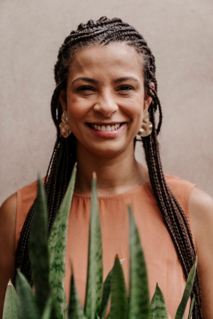 Mulher negra sorrindo para a câmera, com cabelos trançados e blusa terracota, parcialmente enquadrada por folhas verdes em primeiro plano.