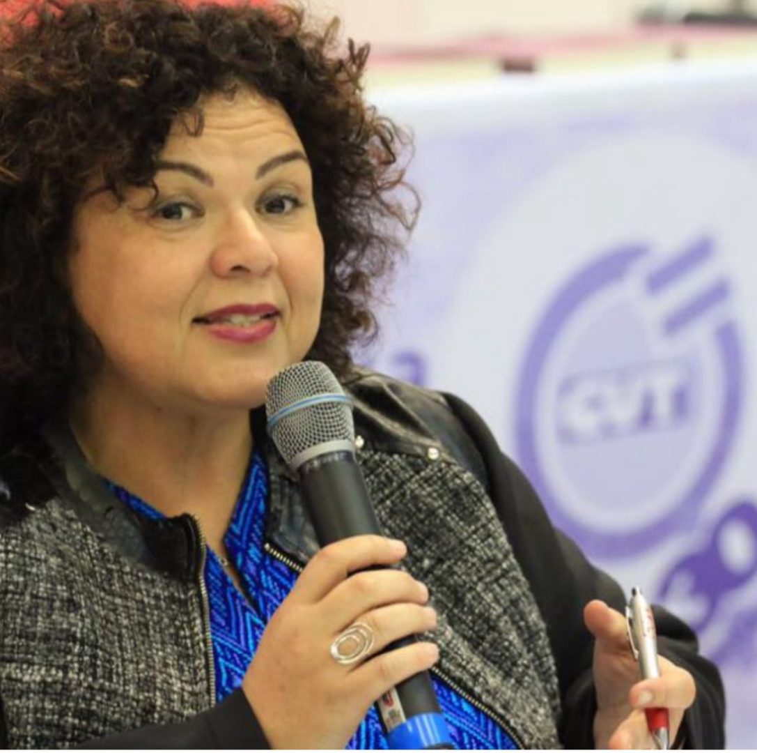 Mulher de cabelos cacheados fala ao microfone durante um evento, segurando uma caneta na outra mão.