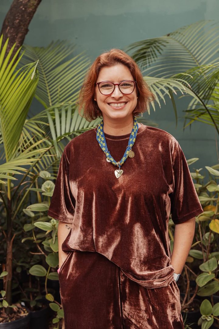 Foto de Mayra Aiello, psicóloga e fundadora do Instituto Doulas de Adoção, em ambiente externo com vegetação ao fundo.