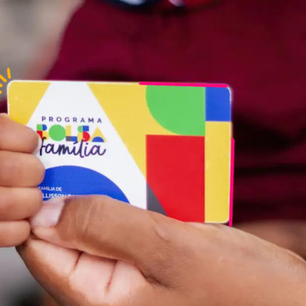 Uma criança segura, com as duas mãos, um cartão do programa Bolsa Família em primeiro plano. O cartão ocupa o centro da imagem, enquanto a criança veste uma camiseta vermelha. O fundo está desfocado, destacando o cartão e as mãos como elementos principais da fotografia.