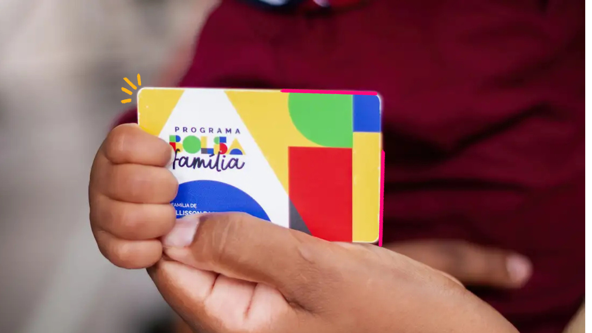 Uma criança segura, com as duas mãos, um cartão do programa Bolsa Família em primeiro plano. O cartão ocupa o centro da imagem, enquanto a criança veste uma camiseta vermelha. O fundo está desfocado, destacando o cartão e as mãos como elementos principais da fotografia.