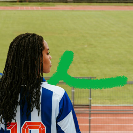 Foto colorida mostra uma menina negra de costas, com longos cabelos pretos trançados. Ela veste uma camisa de futebol azul e branca com o número 10 e olha para o lado. Ao fundo, um campo gramado e uma pista de atletismo.