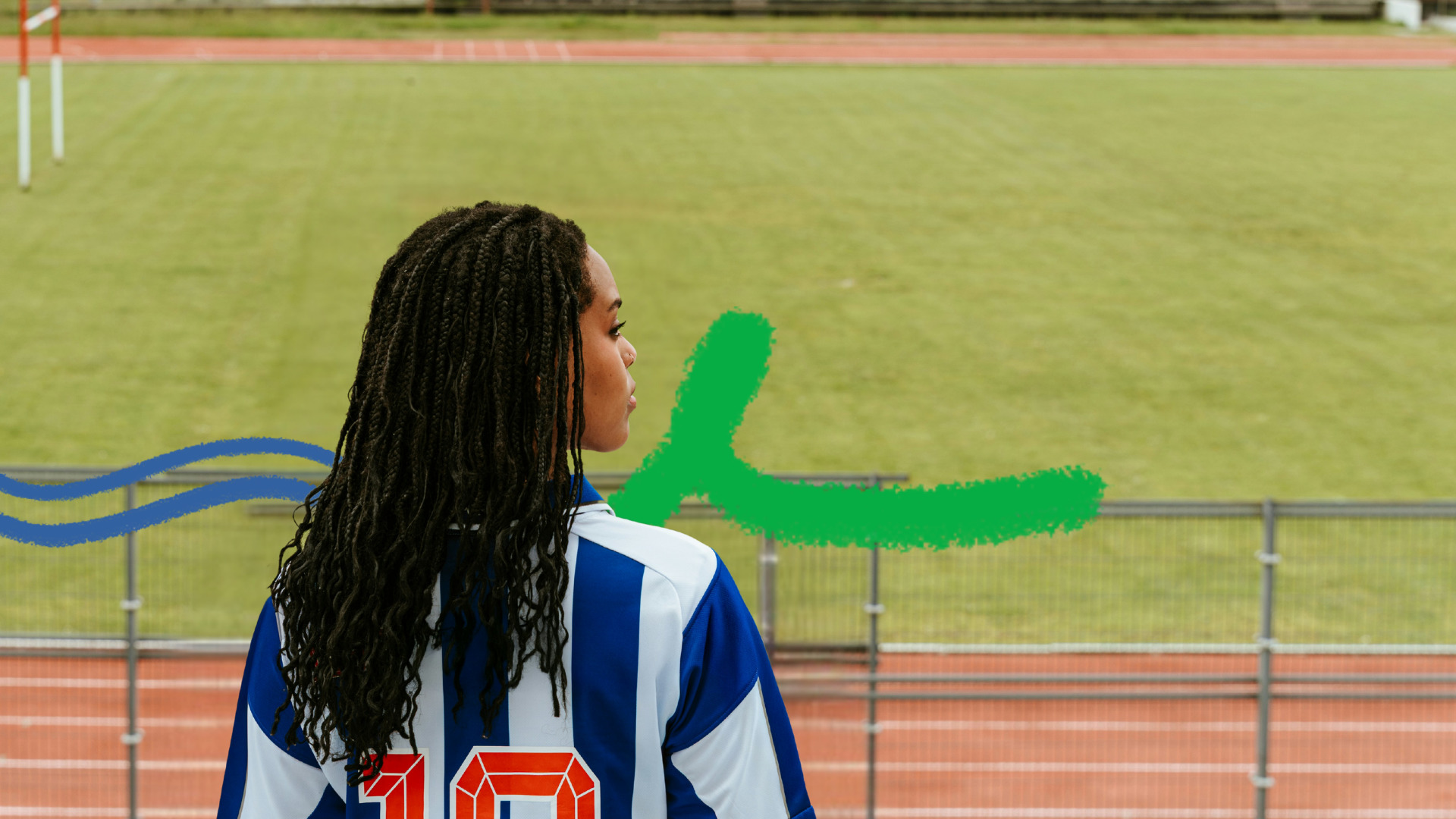 Foto colorida mostra uma menina negra de costas, com longos cabelos pretos trançados. Ela veste uma camisa de futebol azul e branca com o número 10 e olha para o lado. Ao fundo, um campo gramado e uma pista de atletismo.