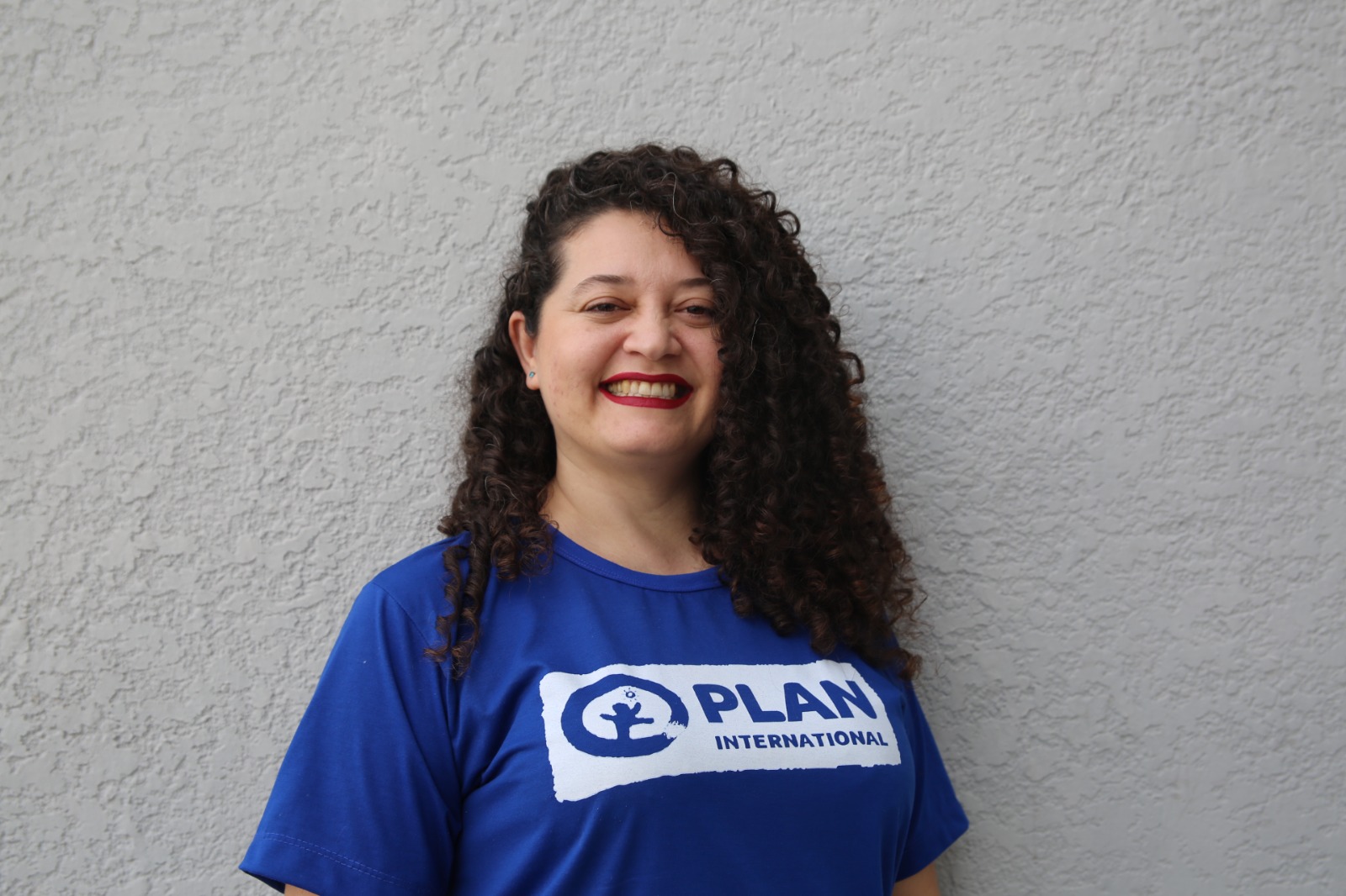 Foto colorida mostra Ana Nery, mulher de cabelos castanhos cacheados, sorrindo. Ela veste uma camiseta azul com o logo da Plan International e está diante de uma parede cinza.