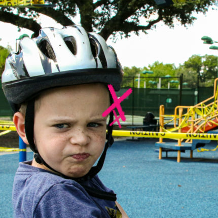 Criança usando capacete faz expressão de recusa em frente a um parquinho interditado com fitas amarelas.