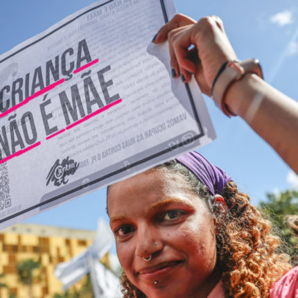 Uma mulher negra em uma manifestação na rua segura um cartaz onde se lê Criança não é mãe em referência a meninas vítimas de estupro