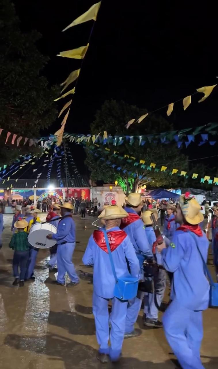 Homens e crianças de uniforme azul e chapéu de palha tocam tambor em praça enfeitada com bandeirinhas, à noite