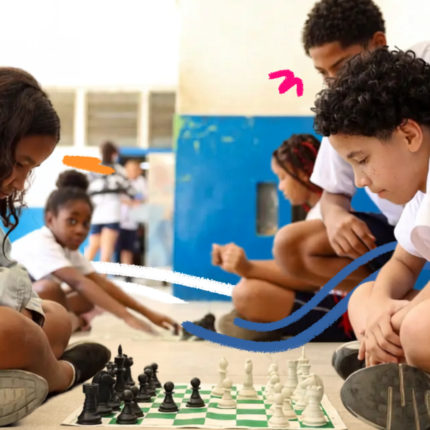 Crianças e adolescentes sentados no chão de uma escola jogam xadrez em pequenos grupos. Ao fundo, outros estudantes conversam e brincam. A imagem recebe elementos gráficos coloridos sobrepostos e ilustra o debate sobre a lei do celular nas escolas e seus impactos.