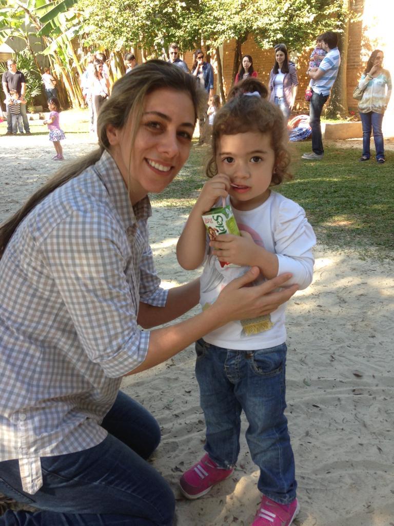 Mulher agachada ao lado de uma menina, que segura uma caixinha de bebida com um canudo. As duas estão em uma área externa, com árvores, plantas e outras pessoas ao fundo.