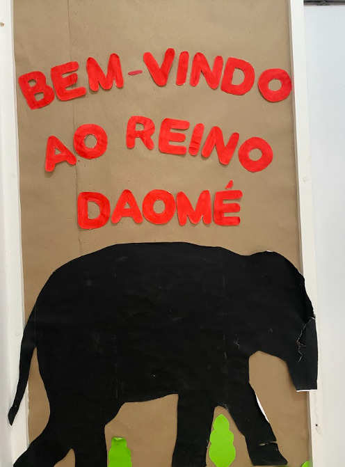 Cartaz de papelão com letras vermelhas escrito “Bem-vindo ao reino daomé”, acompanhado da imagem de um animal preto semelhante a um leão.