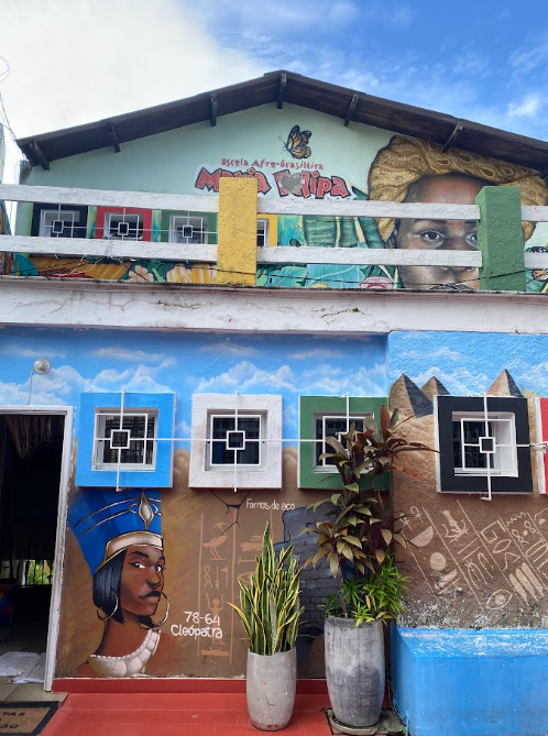 Fachada da Escola Afro-brasileira Maria Felipa, com paredes coloridas e murais que retratam pessoas negras e referências africanas. Na parte superior, há um mural com o rosto de uma pessoa negra e, na parte inferior, uma representação de Cleópatra, além de plantas e janelas.