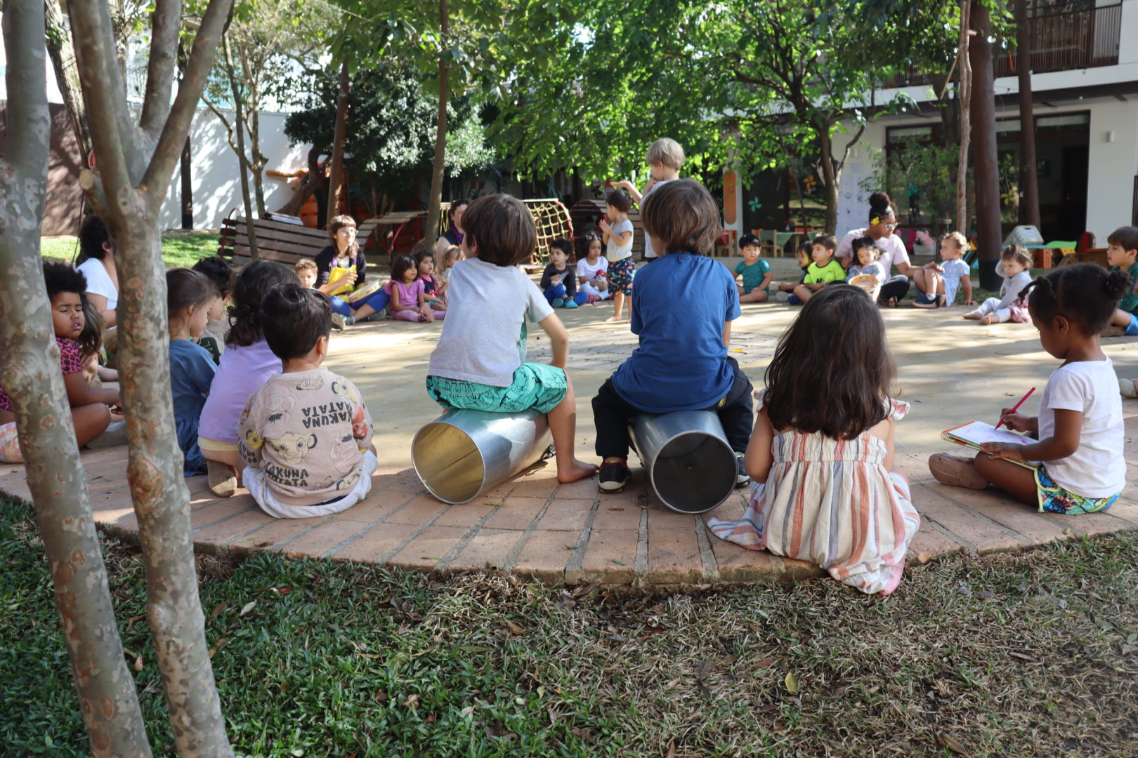 Crianças sentadas em círculo em um espaço externo arborizado, durante uma atividade ao ar livre. Algumas estão sentadas em brinquedos de metal, enquanto outras acompanham a atividade no chão. Ao fundo, há árvores, bancos, brinquedos e o prédio da escola.