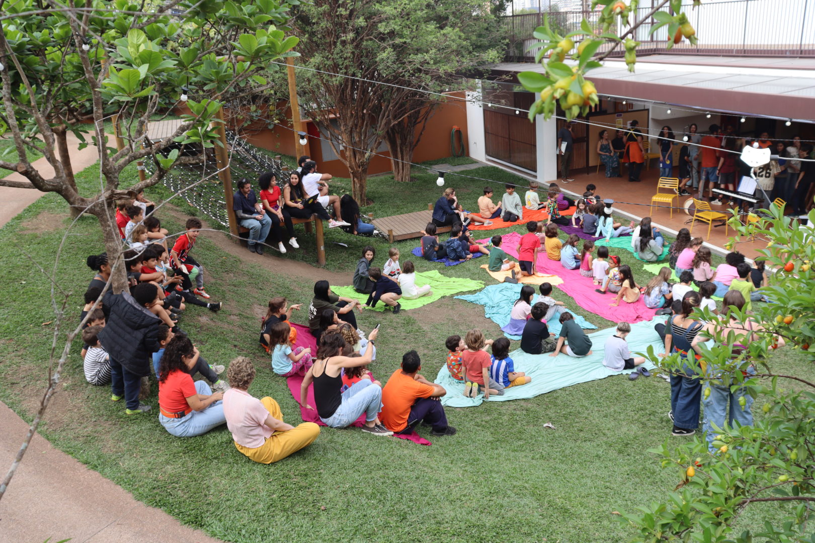Crianças e adultos participam de uma atividade ao ar livre no jardim de uma escola. As crianças estão sentadas sobre tecidos coloridos no gramado, enquanto adultos acompanham a atividade.