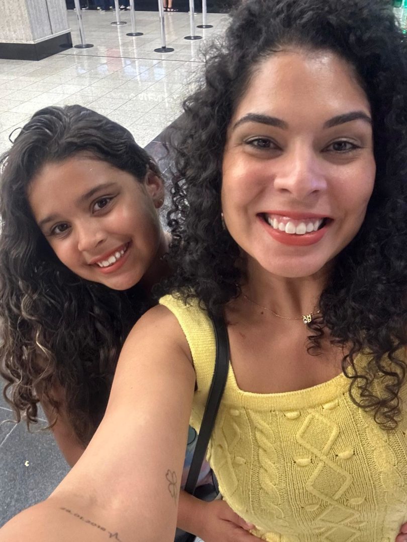 Mulher e menina de cabelos cacheados aparecem sorrindo para a câmera em uma selfie. As duas estão em um ambiente interno, com piso claro e filas de espera ao fundo.