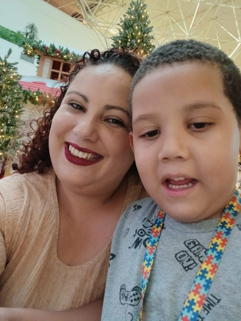 Mulher e menino aparecem juntos em uma selfie. Ela sorri para a câmera, enquanto ele olha para frente. Ao fundo, há árvores de Natal iluminadas e decoração natalina em um ambiente interno.