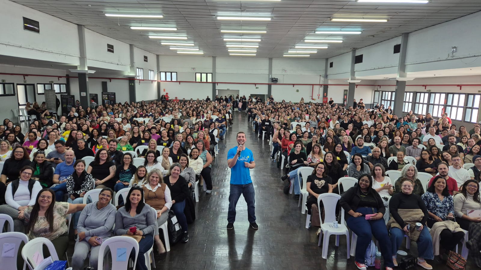 Auditório amplo e lotado, com centenas de pessoas sentadas em fileiras de cadeiras. Um homem de camiseta azul segura um microfone no centro do espaço, diante do público.