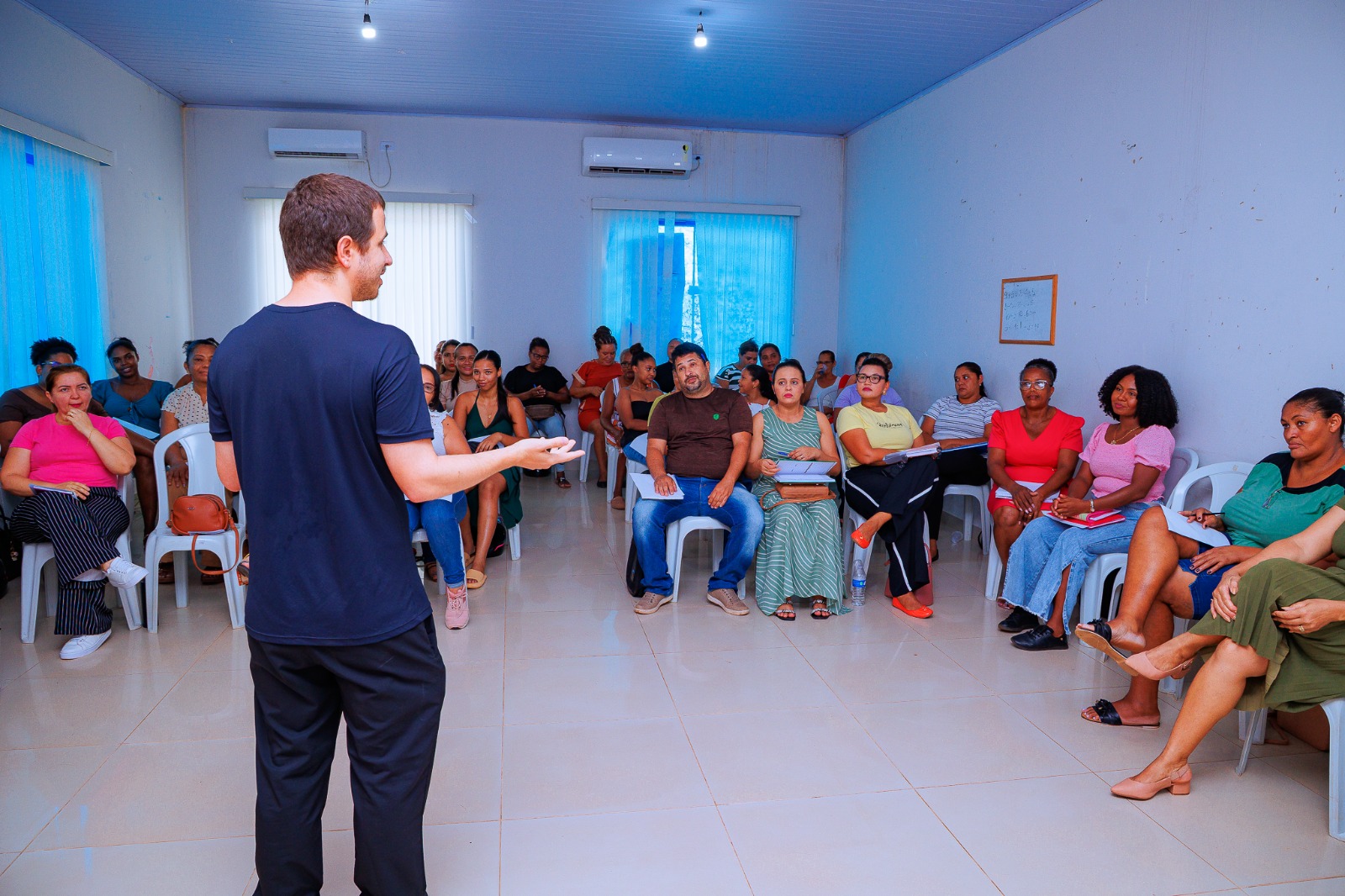 Homem de costas para a câmera fala para um grupo de adultos sentados em círculo em uma sala. Os participantes observam o palestrante, alguns com cadernos nas mãos.