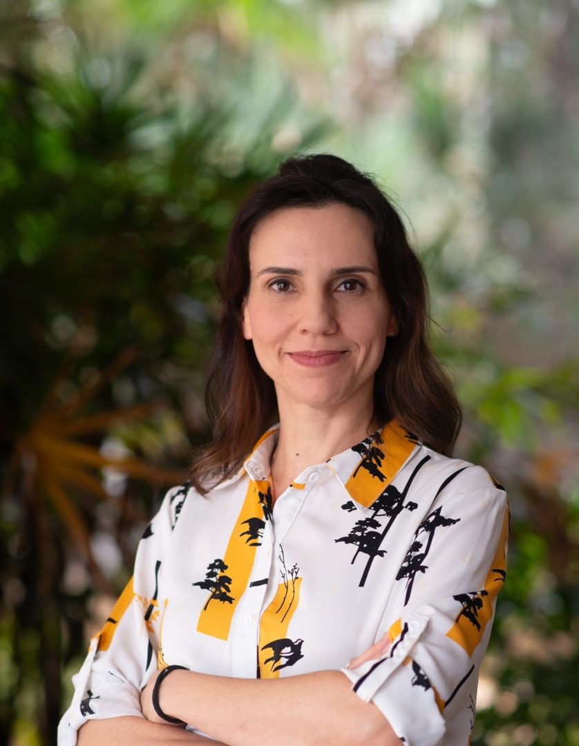 Retrato de Anna Cunha, em frente a uma área verde, usando camisa branca estampada.