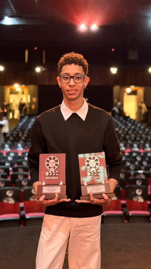 Arthur Campos, 16 anos, segura dois troféus em um auditório com cadeiras vermelhas.
