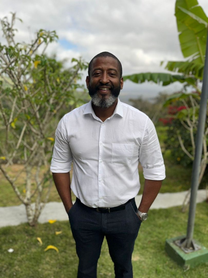 Homem negro, de barba grisalha, usando camisa branca e calça escura, posa sorrindo em uma área externa com árvores ao fundo.