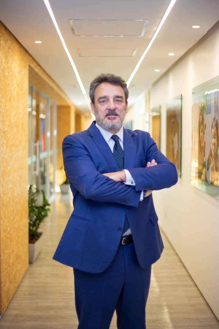 Homem de terno azul posa com os braços cruzados em um corredor de escritório.