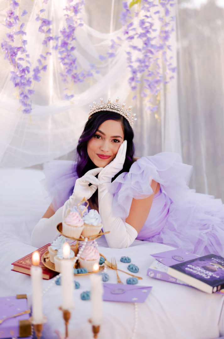 Adolescente deitada sobre uma cama branca, usando vestido lilás, luvas brancas e tiara. Ela sorri para a câmera, apoiando o rosto nas mãos. Ao redor, há livros, cupcakes, velas acesas, cartões e flores em tons de lilás.