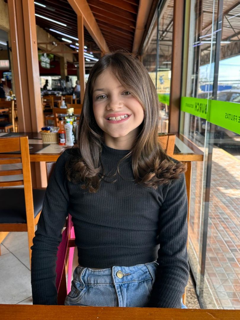 Manuela Malavazi, 9 anos, sentada em uma mesa de restaurante e sorrindo para a câmera.