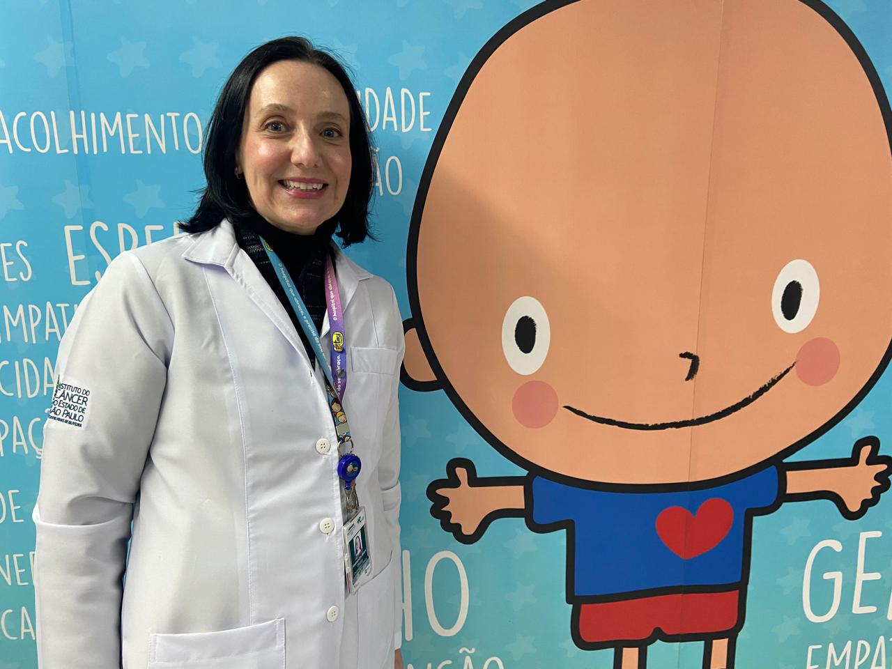 Mulher de cabelos pretos, usando jaleco branco e crachá, sorri ao lado de um painel azul com a ilustração de uma criança.