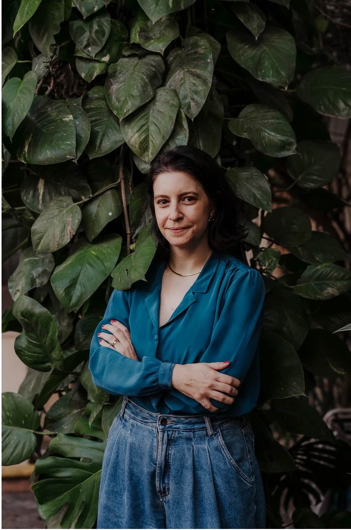 Mulher de óculos, blusa azul e calça jeans posa sorrindo, com os braços cruzados, diante de uma parede com plantas verdes.