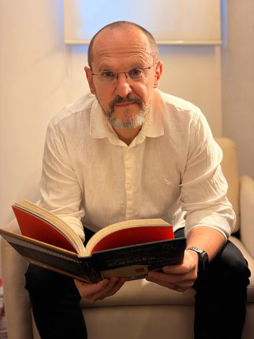 Ilan Brenman, de camisa branca e óculos, segura um livro aberto enquanto está sentado em uma poltrona.