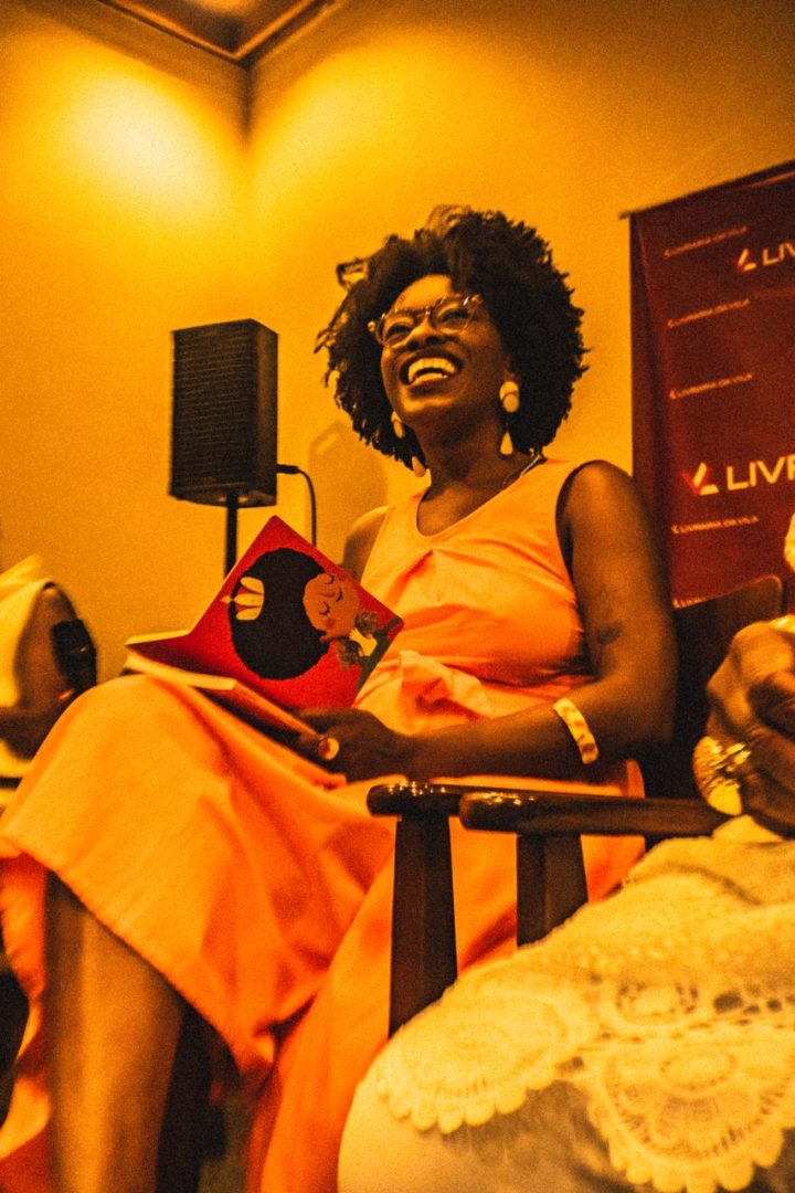 Jussara Santos sorri durante um evento, sentada em uma cadeira e segurando um livro ilustrado.