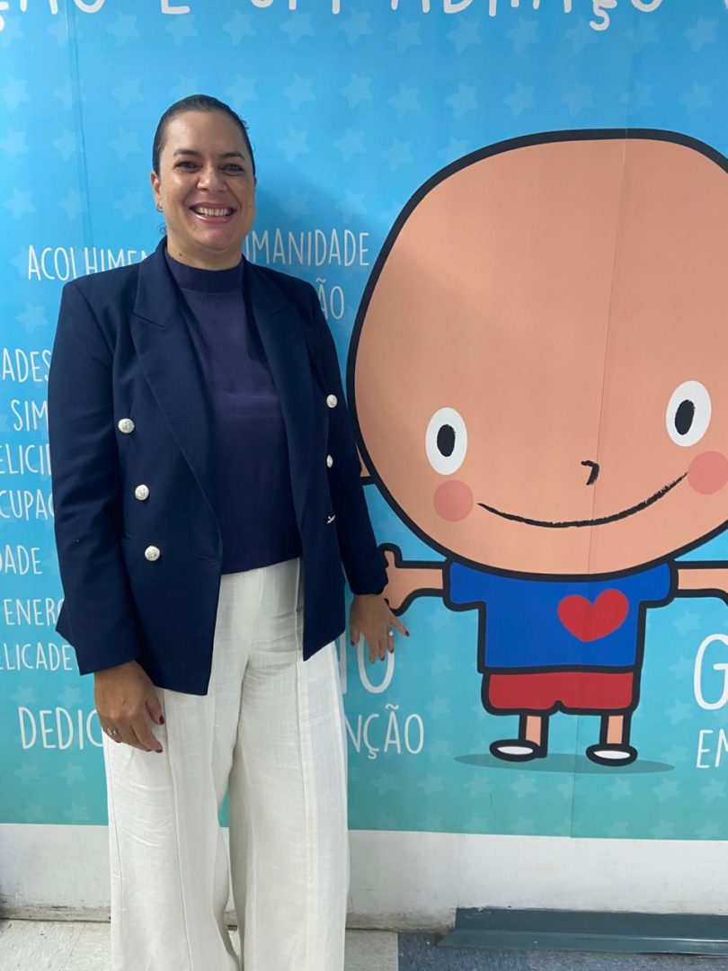 Mulher de blazer azul-marinho e calça branca sorri para a câmera ao lado de um painel azul com a ilustração de uma criança.
