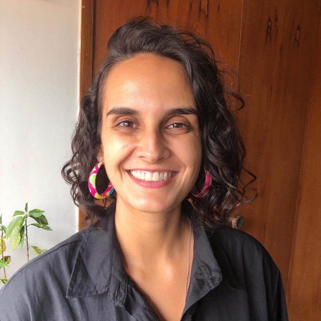 Livia Ribeiro, sócia-fundadora da Reconectta, sorri para a câmera. Ela tem cabelos cacheados, usa brincos coloridos e uma camisa preta.