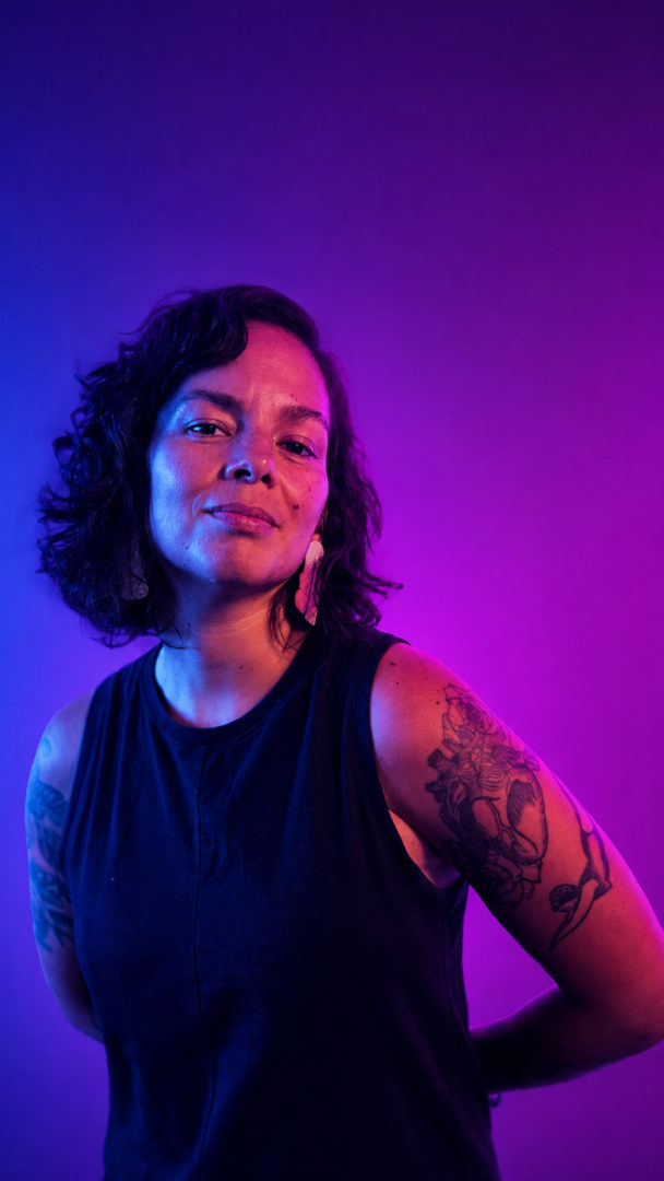Lua Barros posa para um retrato com blusa preta sem mangas, cabelos cacheados curtos, brincos e tatuagens nos braços, sob iluminação azul e rosa.