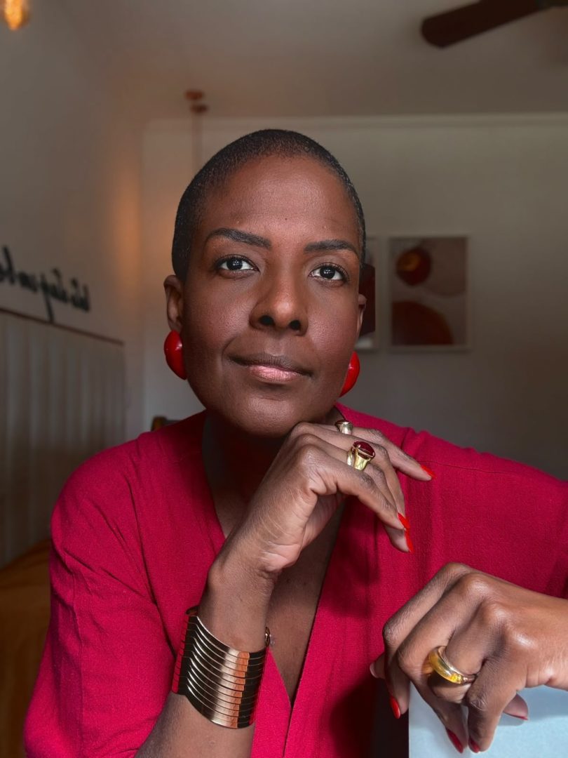 Mulher negra de cabelos curtos usa blusa vermelha, brincos grandes e acessórios dourados. Ela apoia uma das mãos no rosto e olha para a câmera.