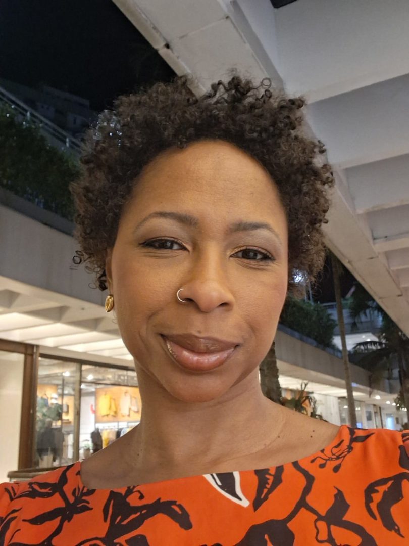 Mulher negra de cabelos curtos e cacheados, com piercing no nariz e blusa laranja estampada, fotografada em um ambiente interno.