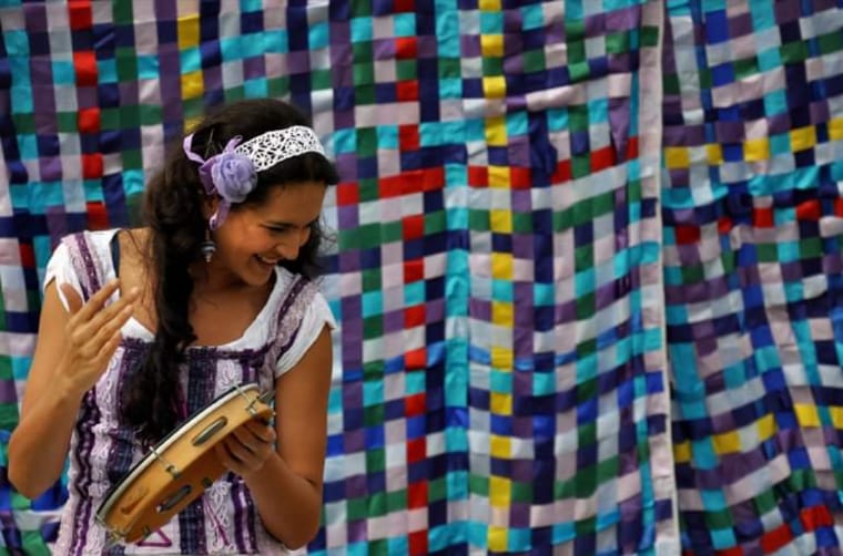 Arte educadora Sabryna Mato Grosso toca pandeiro durante uma atividade teatral, diante de um painel colorido de tecidos.